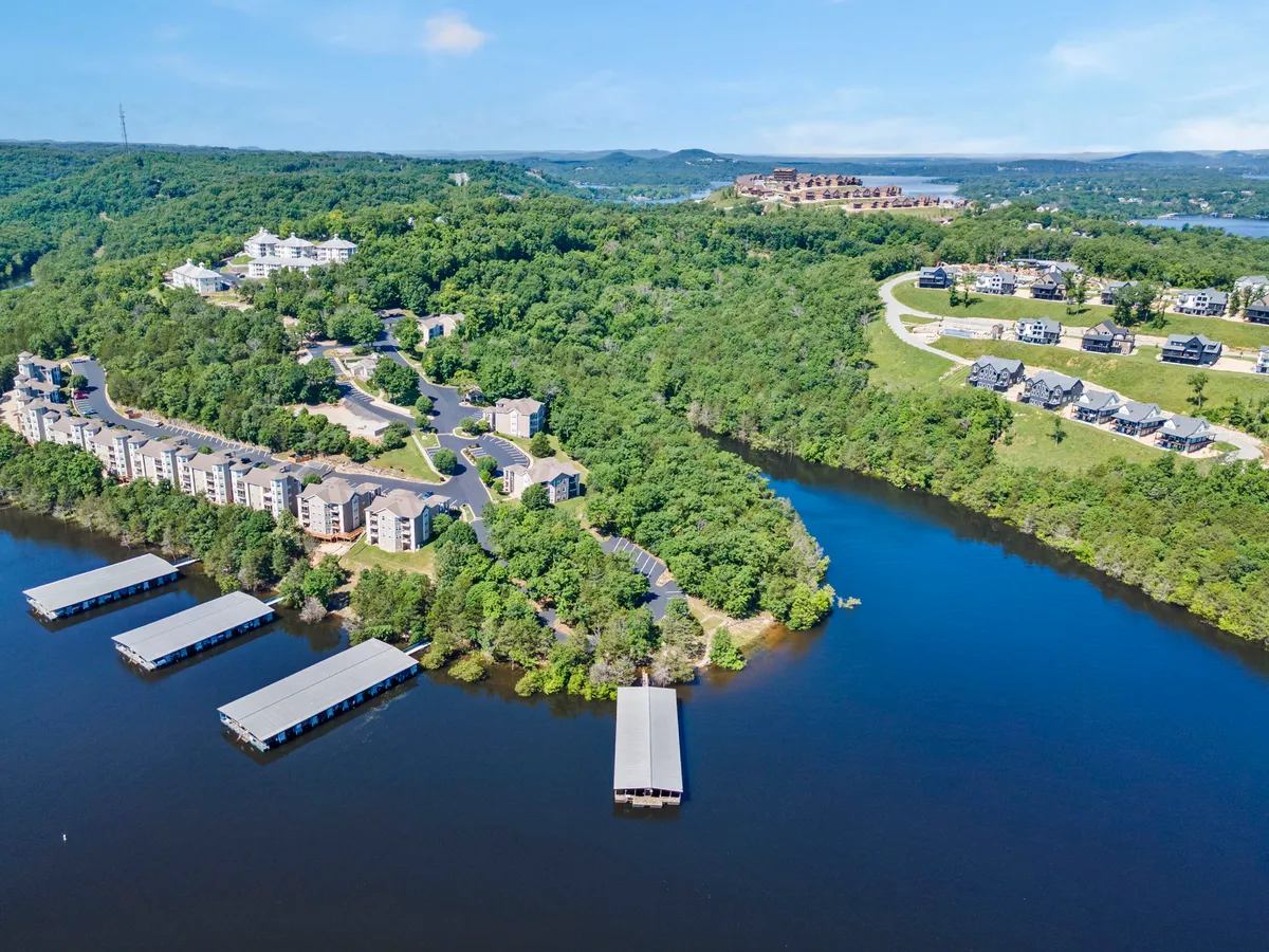 A vibrant scene showing waterfront docks, lush greenery, residential buildings nestled among the trees, and winding roads leading to the lake. The setting is ideal for boating, fishing, hiking, and family gatherings, reflecting both adventure and community spirit.