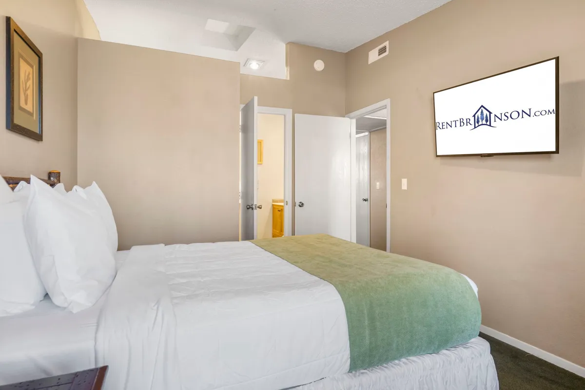 A cozy bedroom featuring a bed with white linens and a green throw, a wall-mounted television, and open doorways that lead into other parts of the suite. The room is neatly organized, welcoming, and filled with soft natural light.