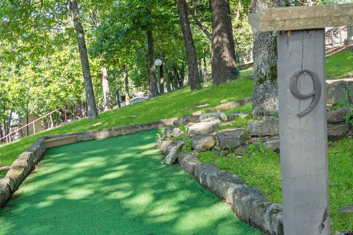 A vibrant mini-golf course set amid natural stone borders and lush greenery, with a wooden post marking the number 9. The scene is playful and inviting, capturing moments of family fun and friendly competition.
