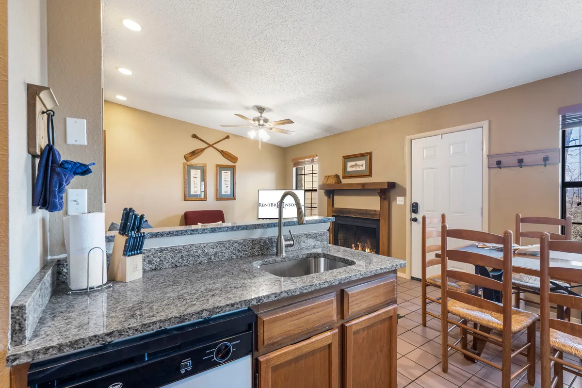 A welcoming open-concept living and dining area featuring a granite countertop, dining table, cozy couch, and a stone fireplace. Sunlight pours through the windows, brightening the space and highlighting the warm, homey atmosphere. The fireplace is a central feature, complemented by plush seating and a seamless flow between the living and dining zones.