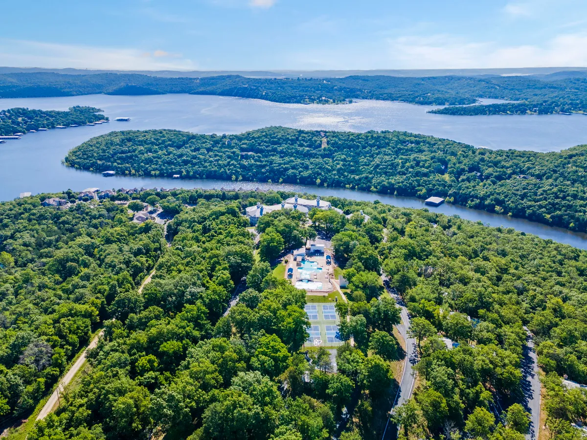 A sweeping view over lush greenery reveals a sparkling swimming pool, tennis courts, and a breathtaking waterfront, all set against rolling hills. The image exudes a sense of resort-style leisure and offers multiple outdoor activities within a peaceful natural landscape.