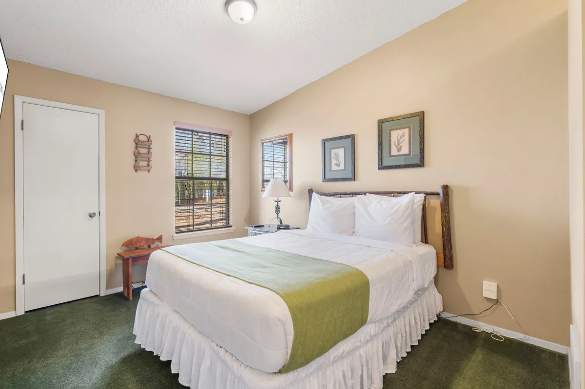 A tranquil bedroom scene featuring a bed with crisp white linens and a green accent throw, complemented by a wooden bedside lamp and framed artwork on the wall. Natural light streams in from a window, creating a soothing and restful ambiance.