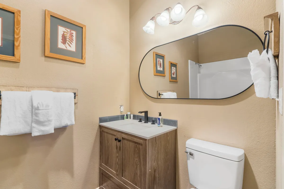 A stylish ensuite bathroom with an oval mirror, wooden vanity, white towels, and framed artwork. The space is bathed in inviting light, suggesting a fresh and comforting self-care experience.