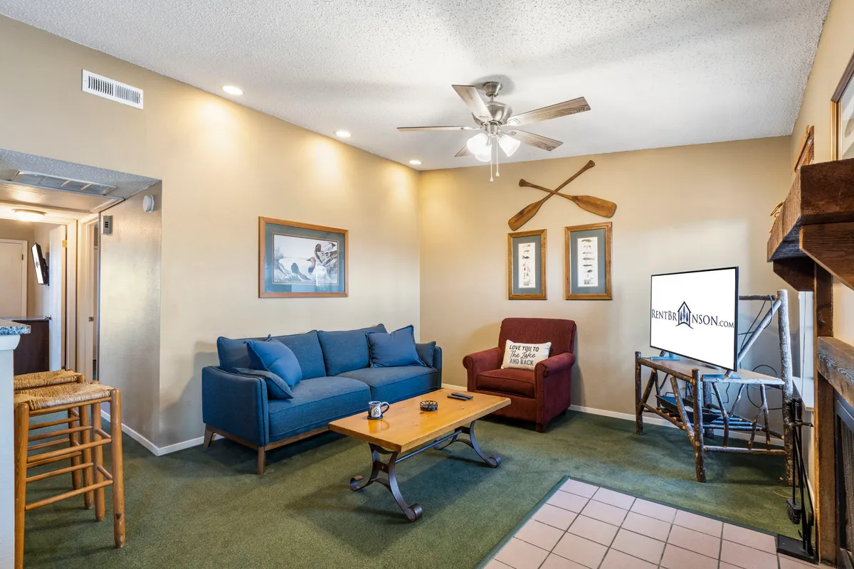 A cozy living room with a blue sofa, wooden coffee table, TV, framed artwork, and a brown armchair. The space is styled for comfort, inviting guests to relax, enjoy movie nights, or gather together for games and conversation.