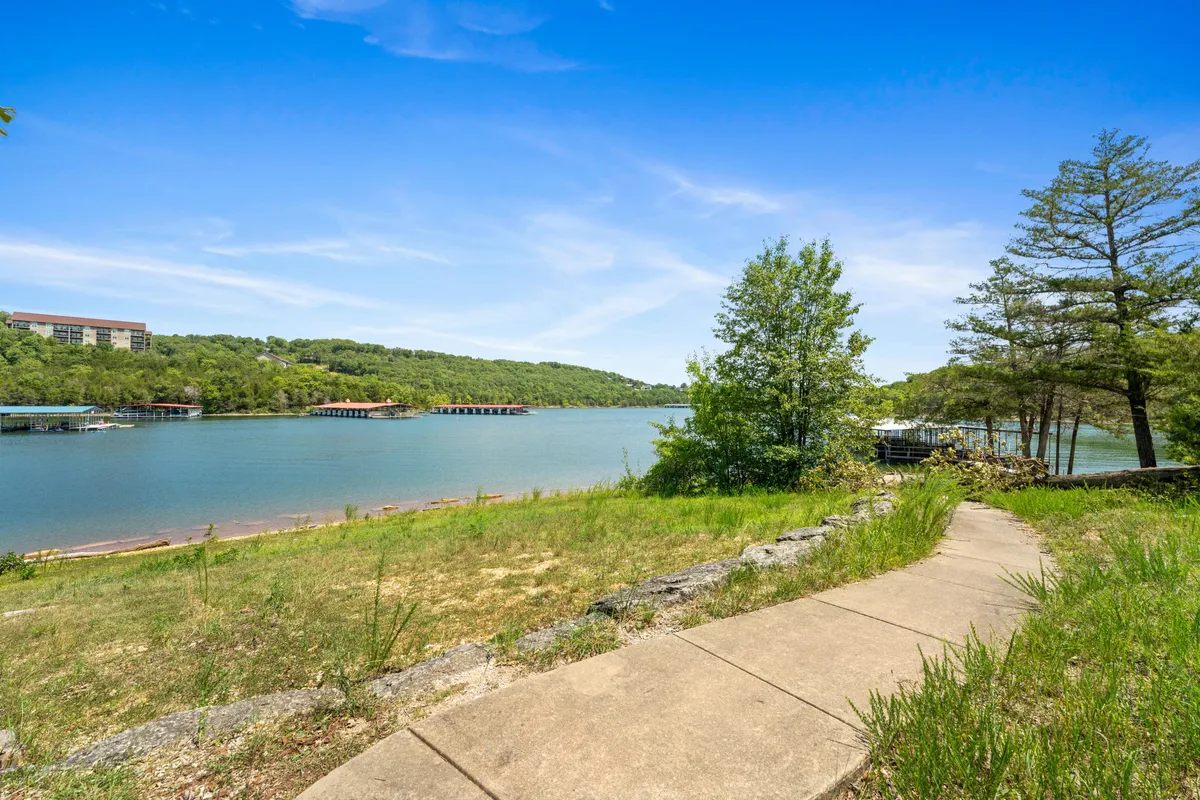 A tranquil view of Table Rock Lake, framed by a pathway, greenery, gentle hills, and several boathouses dotting the shore. The scene captures the calm waters and the lush surroundings, inviting visitors to explore, fish, or relax by the lake.